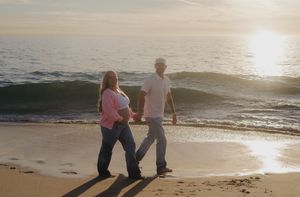 Pregnant couple holding hands strolling on beach at golden sunset with ocean waves.