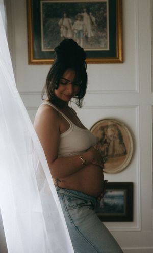 A pregnant woman in a white crop top and jeans cradles her baby bump near sheer curtains.