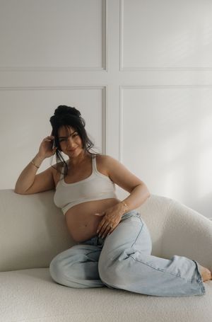 A smiling pregnant woman in a white crop top and jeans relaxes on a white sofa.