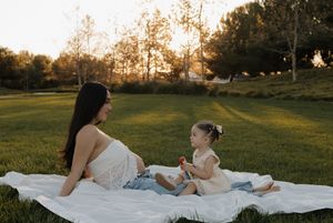 Pregnant mother in white lace top sits on blanket in sunny park sharing a moment with her toddler daughter.
