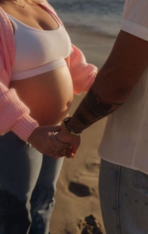 Close-up of tattooed man holding hands with pregnant woman in white crop top on beach.