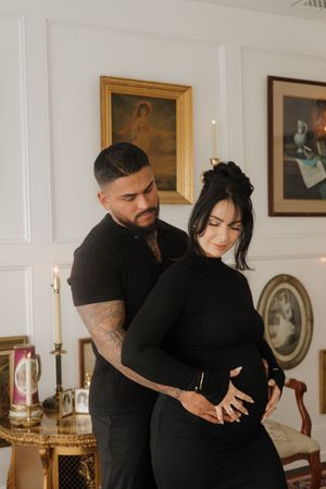 A couple in black outfits tenderly cradles a pregnant belly in an elegant decorated room.