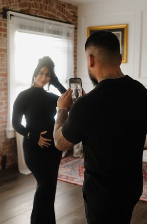 A tattooed man photographs his pregnant partner in a black dress near a sunlit window.
