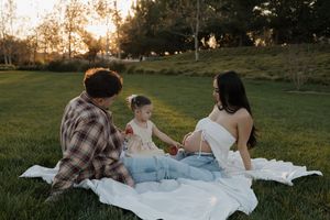 Family of three sits on white blanket in golden sunlight, with pregnant mom, dad, and young toddler daughter.