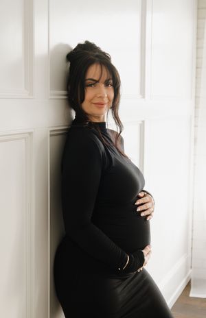 A pregnant woman in a black long-sleeve dress leans against a white wall smiling softly.