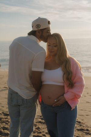 Man kissing pregnant blonde woman on cheek during maternity shoot at sunset beach.
