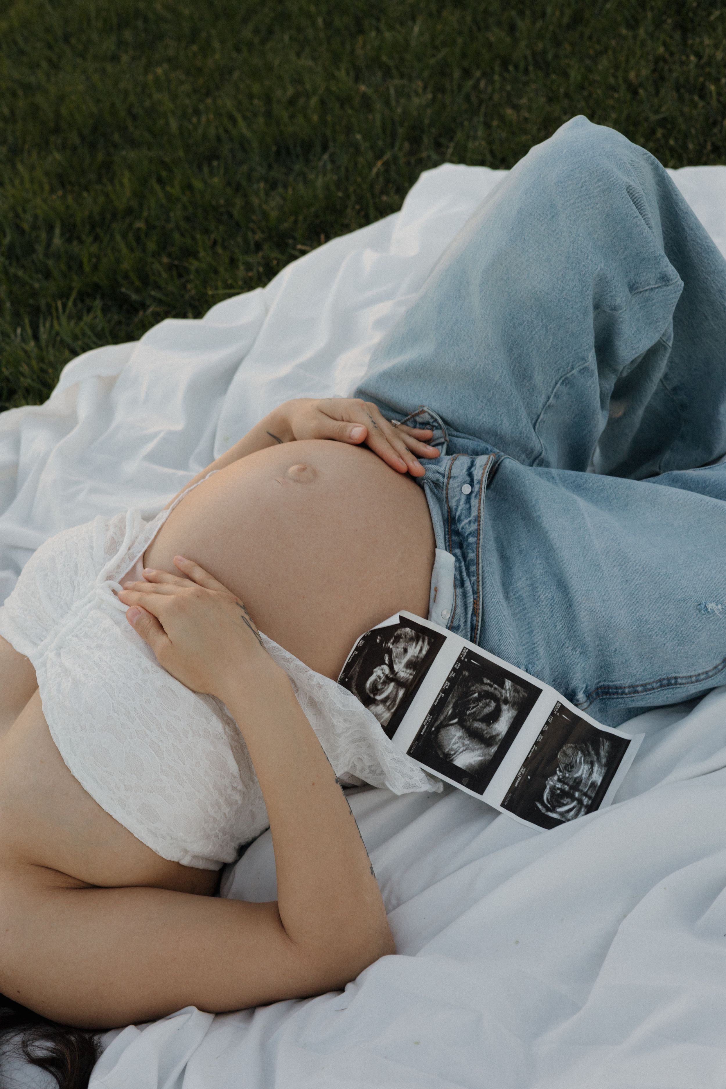 Pregnant woman lies on white blanket outdoors, cradling her bare baby bump with ultrasound photos resting on belly.
