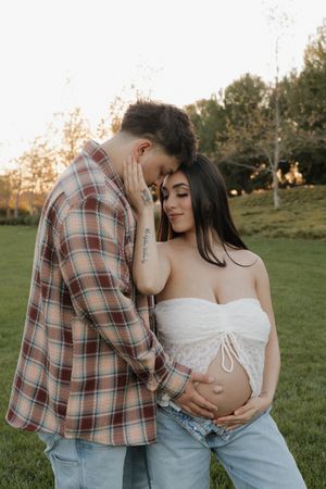 Couple shares tender moment at sunset, with man nuzzling pregnant partners head as she cradles her baby bump.