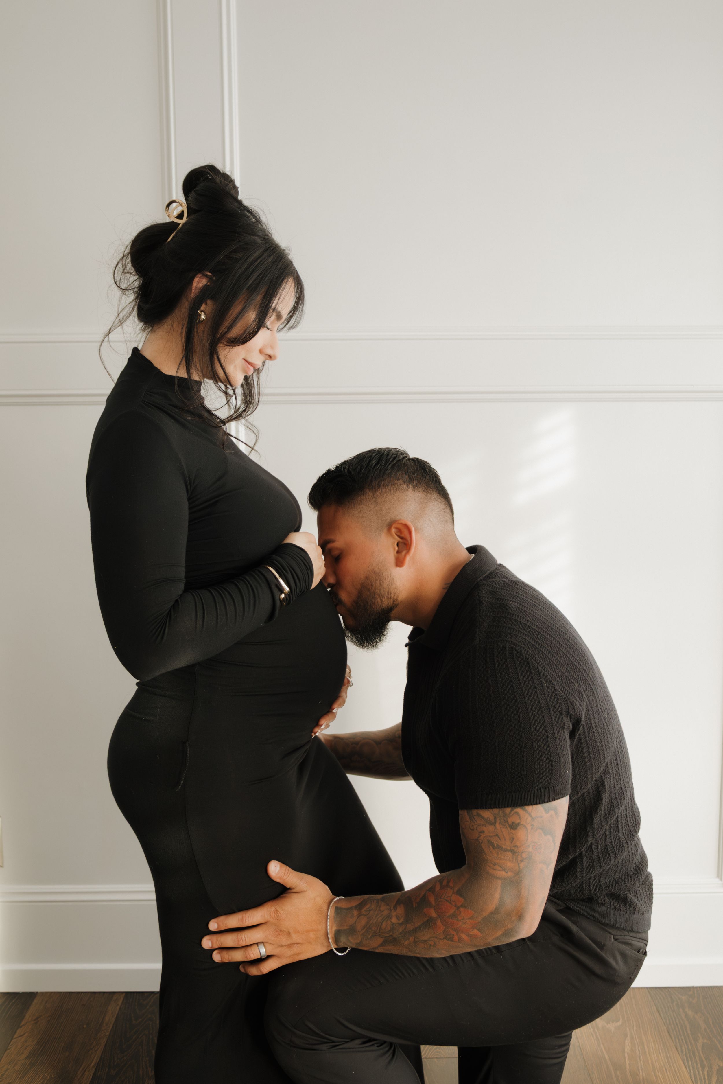 Couple in black outfits during maternity photo shoot, man kissing pregnant woman's belly indoors.