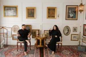Couple dressed in black sitting in elegant room with framed artwork and antique furniture.