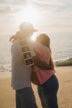 Expecting couple embracing and kissing backlit by golden sunset on beach with ultrasound.