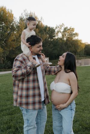 Proud dad lifts toddler on his shoulders while pregnant mom smiles during golden hour family maternity photoshoot.