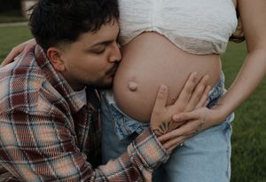 Loving partner kisses pregnant womans bare baby bump while gently holding her during outdoor maternity photoshoot.