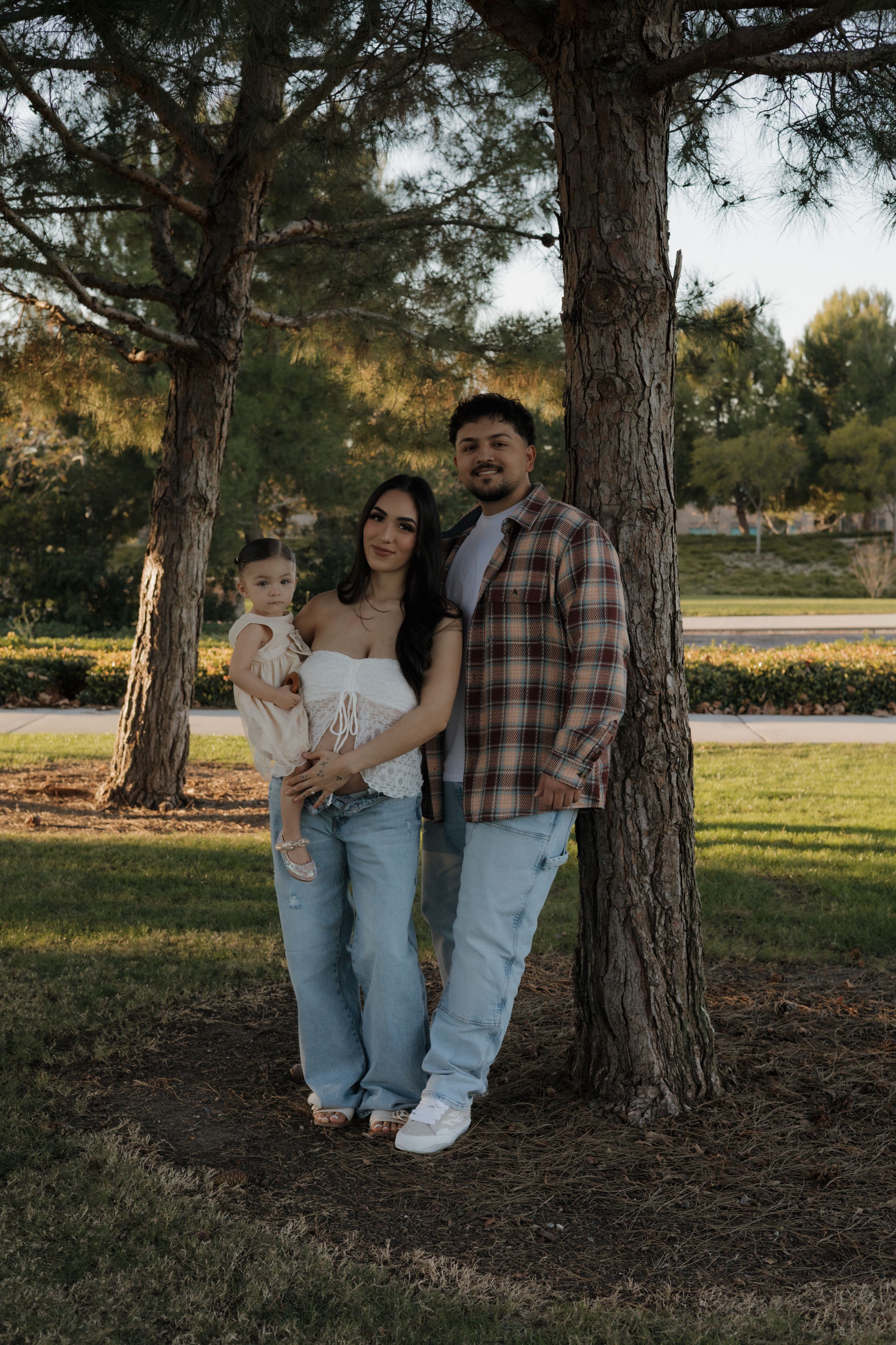 Family of three poses under pine trees in a park during golden hour, parents holding young child.