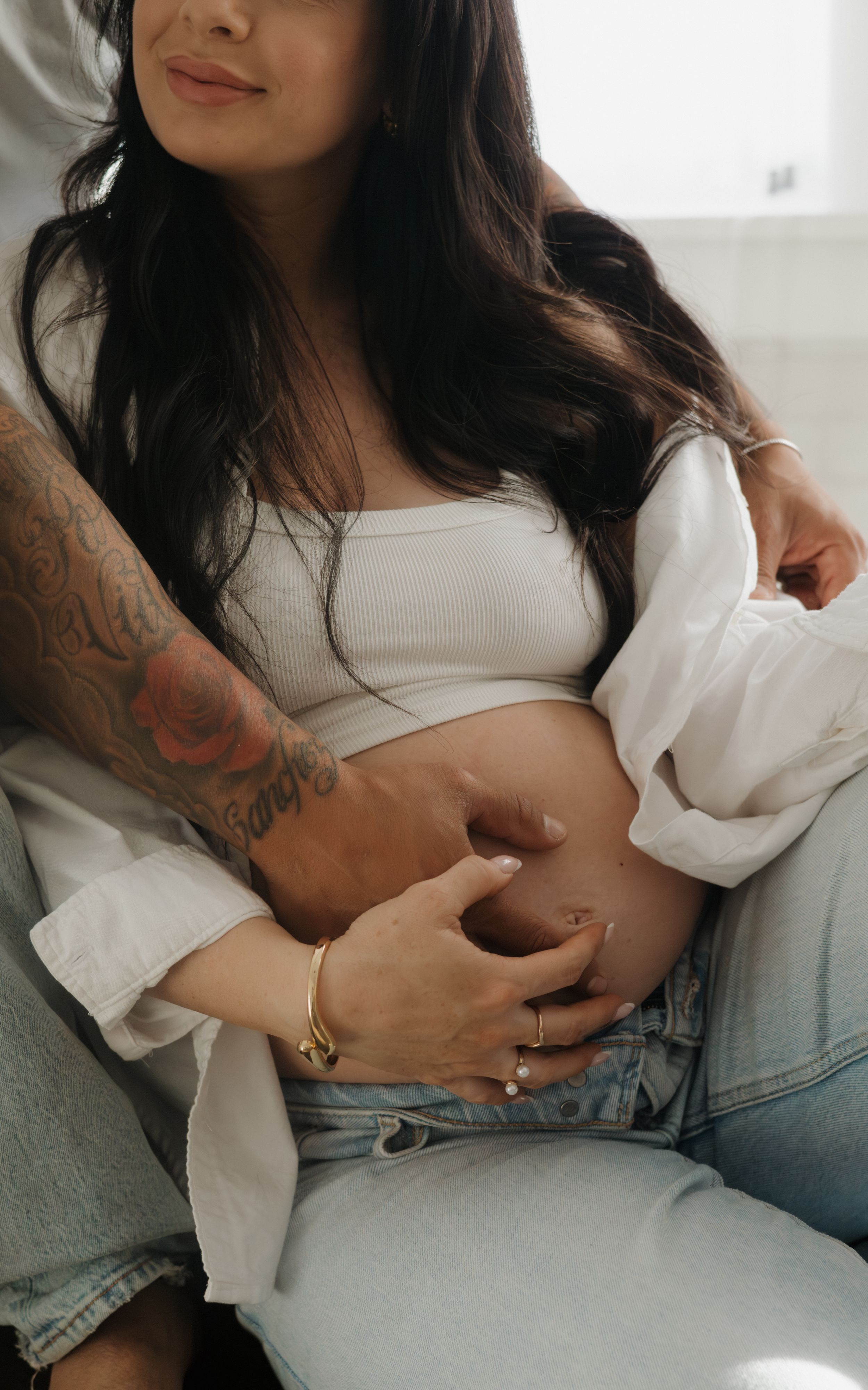 Close-up of a pregnant woman in a white crop top with a tattooed arm embracing her baby bump.