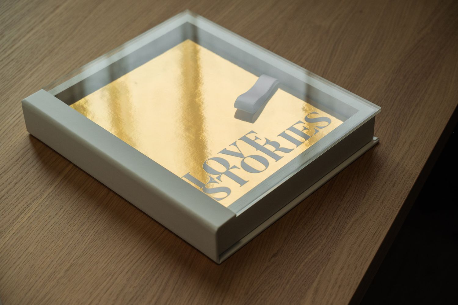 Love Stories gold metallic photo book displayed in white acrylic case on wooden surface.