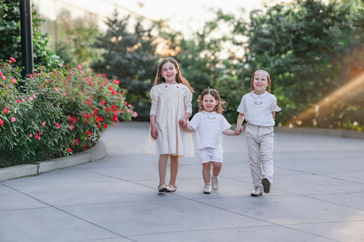 Three children in white outfits walk hand-in-hand along a garden path lined with red flowers.