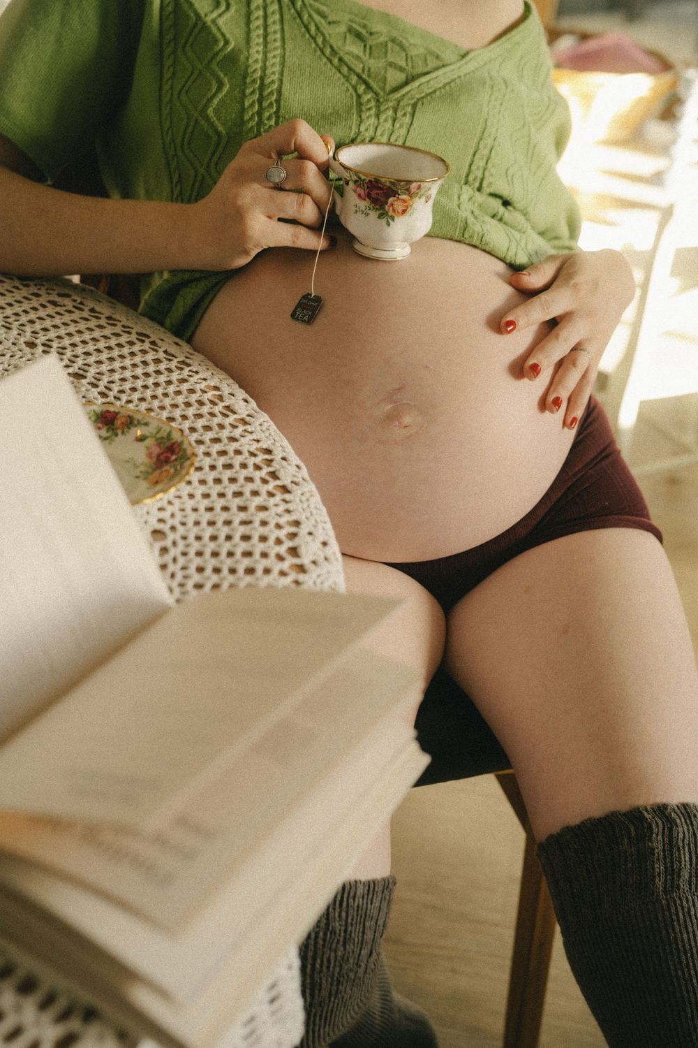 Pregnant woman in green top holding teacup, sitting with a book, wearing knee socks and burgundy shorts.