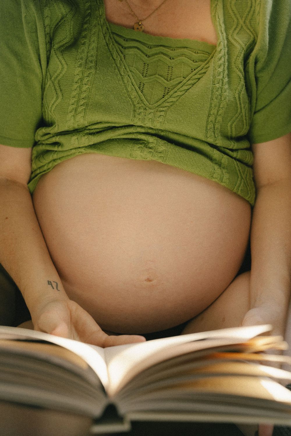 Pregnant woman in green top reading a book, showing bare baby bump, prenatal education concept.