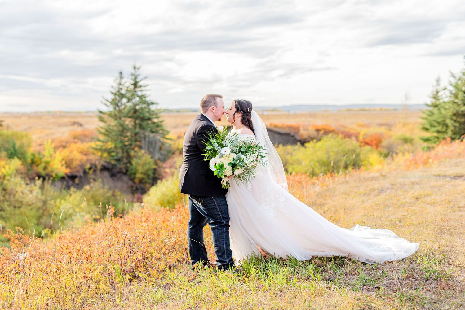 Bride and groom kissing on a hilltop surrounded by vibrant autumn foliage with a bouquet of white flowers.