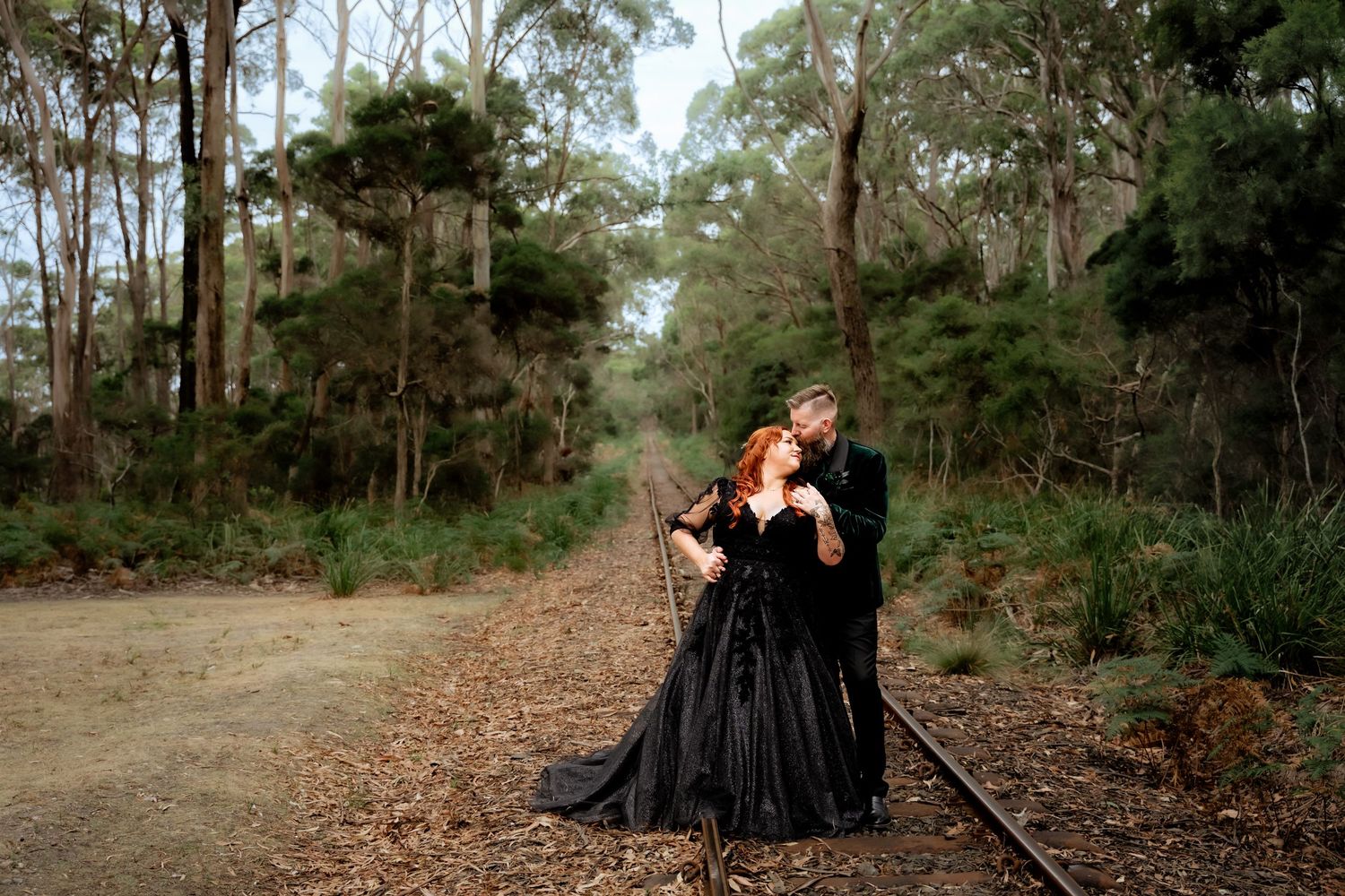 Couple in black gothic wedding attire embracing on railway tracks surrounded by lush forest.