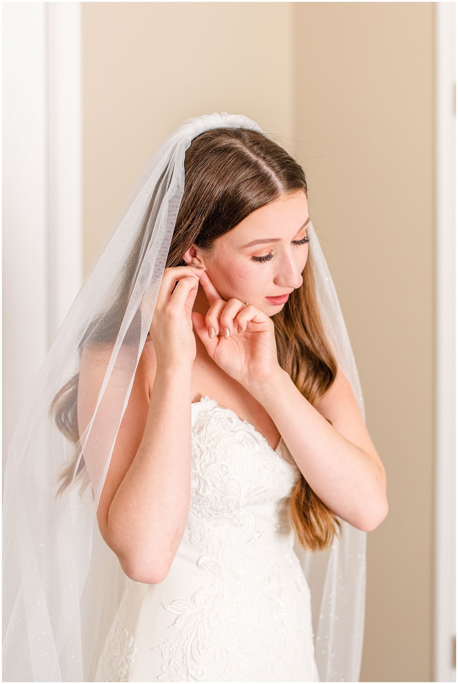 Beautiful bride in strapless lace gown and veil putting on earrings while looking down in soft natural light.
