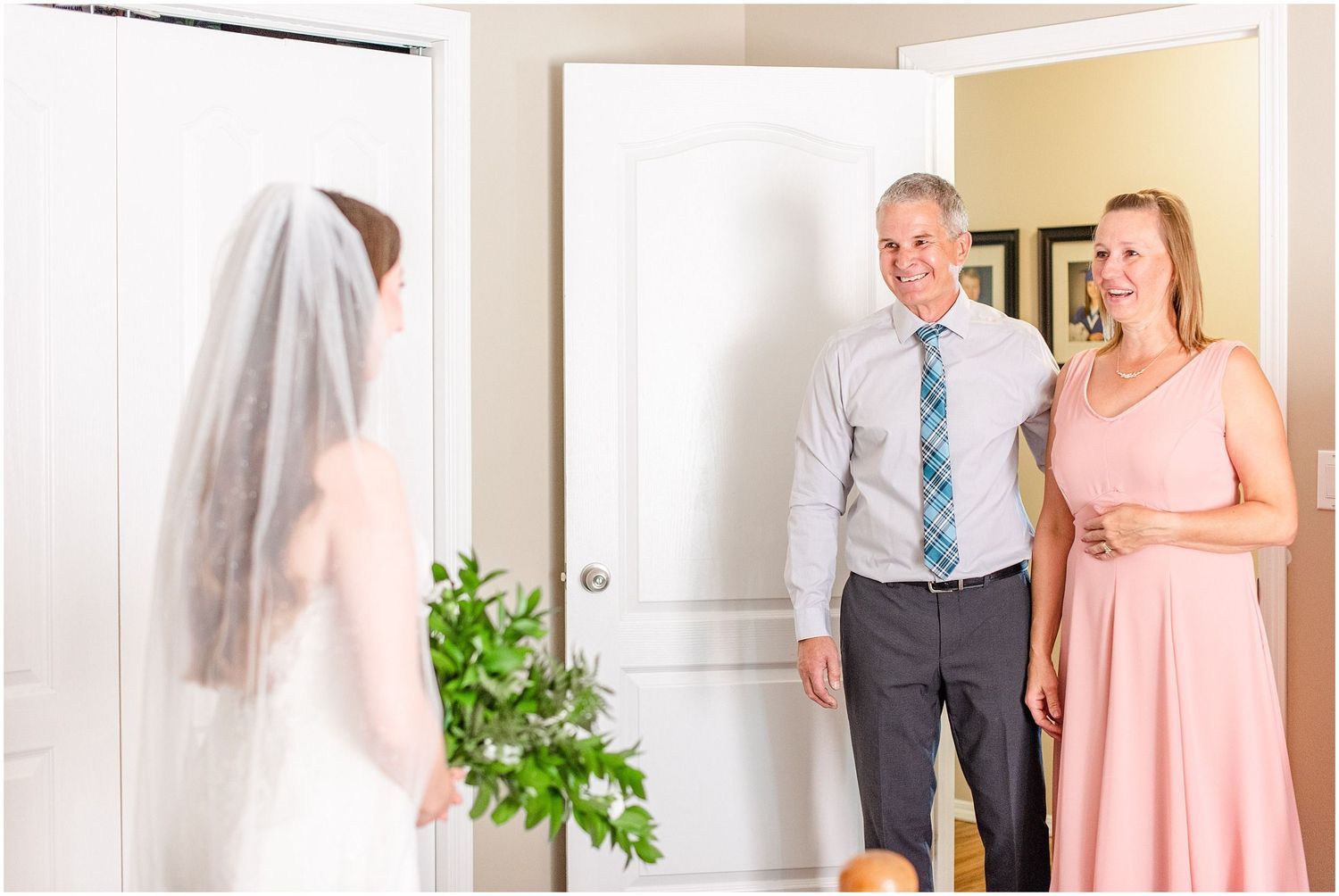 Bride in white gown and veil shows her wedding look to smiling parents in a bright doorway.