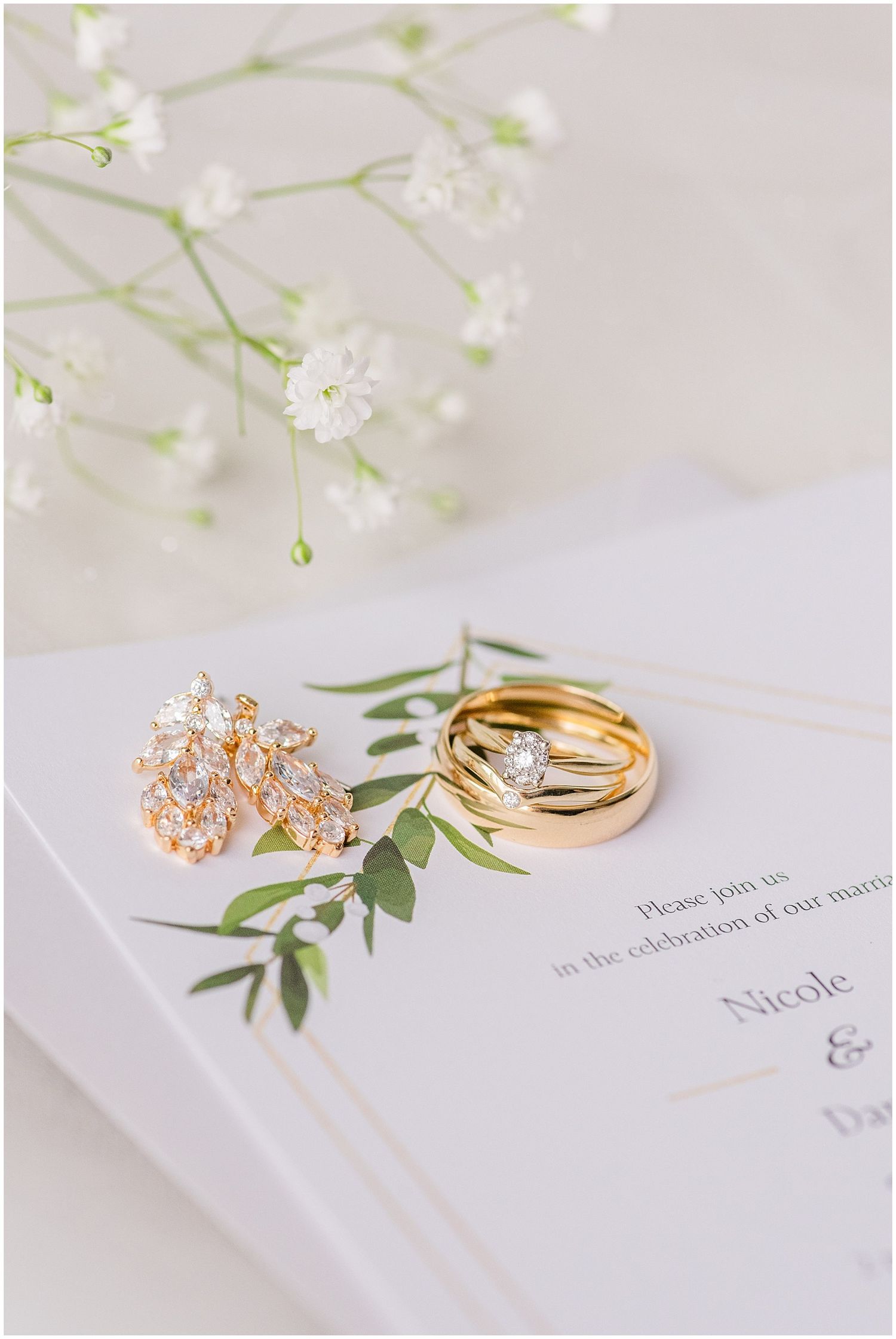 Gold wedding rings, diamond earrings, and greenery on a white wedding invitation with baby's breath flowers.