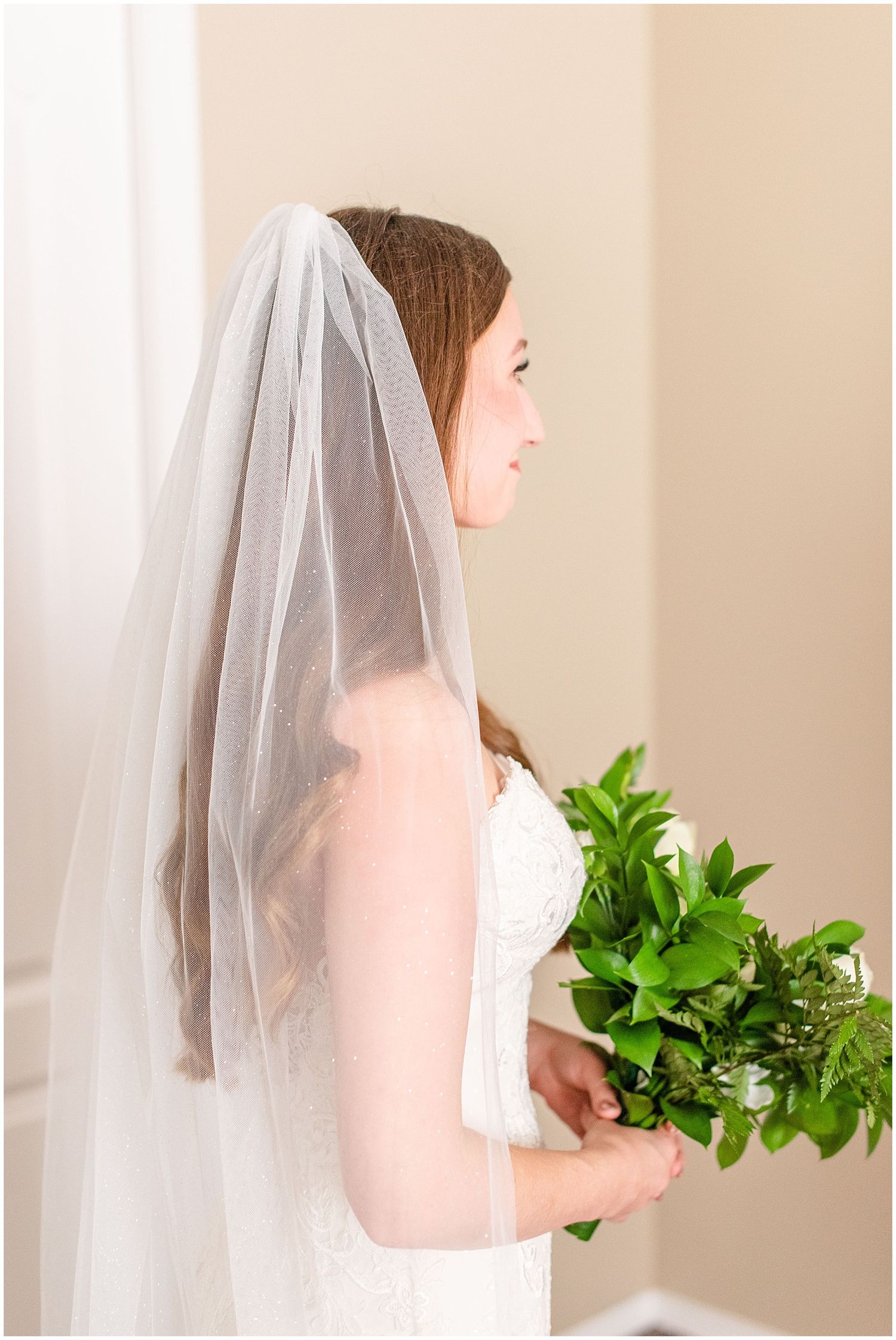 Elegant bride in lace gown and veil holds a green bouquet while posing in soft natural light.