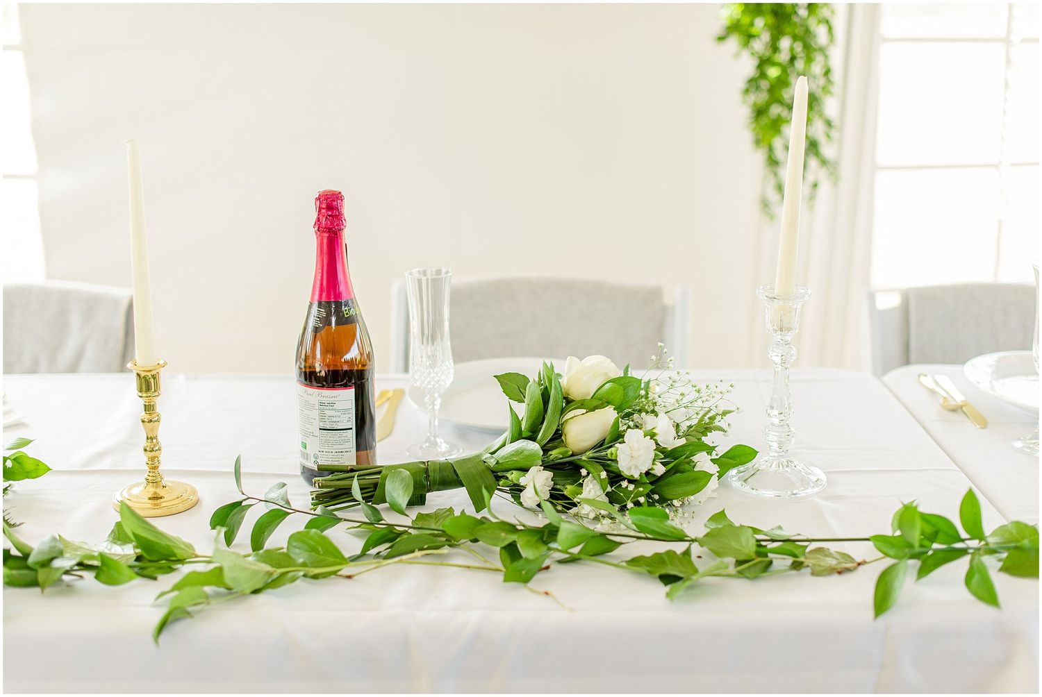 Elegant wedding table with white floral greenery garland, champagne bottle, gold candlestick, and crystal glasses on white linen.