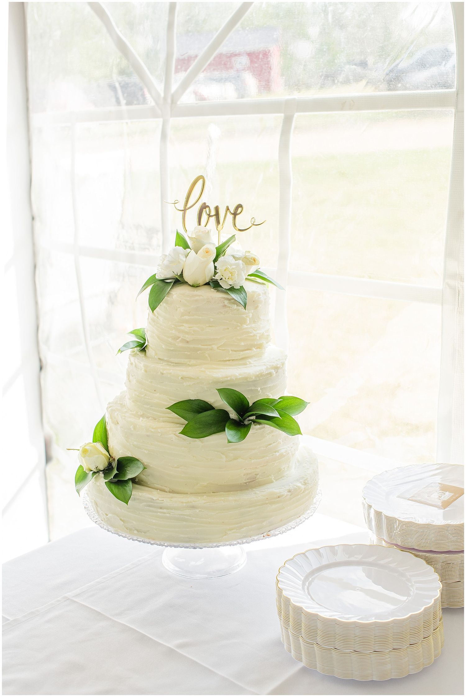 Three-tier white wedding cake with green leaves, white flowers, and gold love topper on white cake stand.