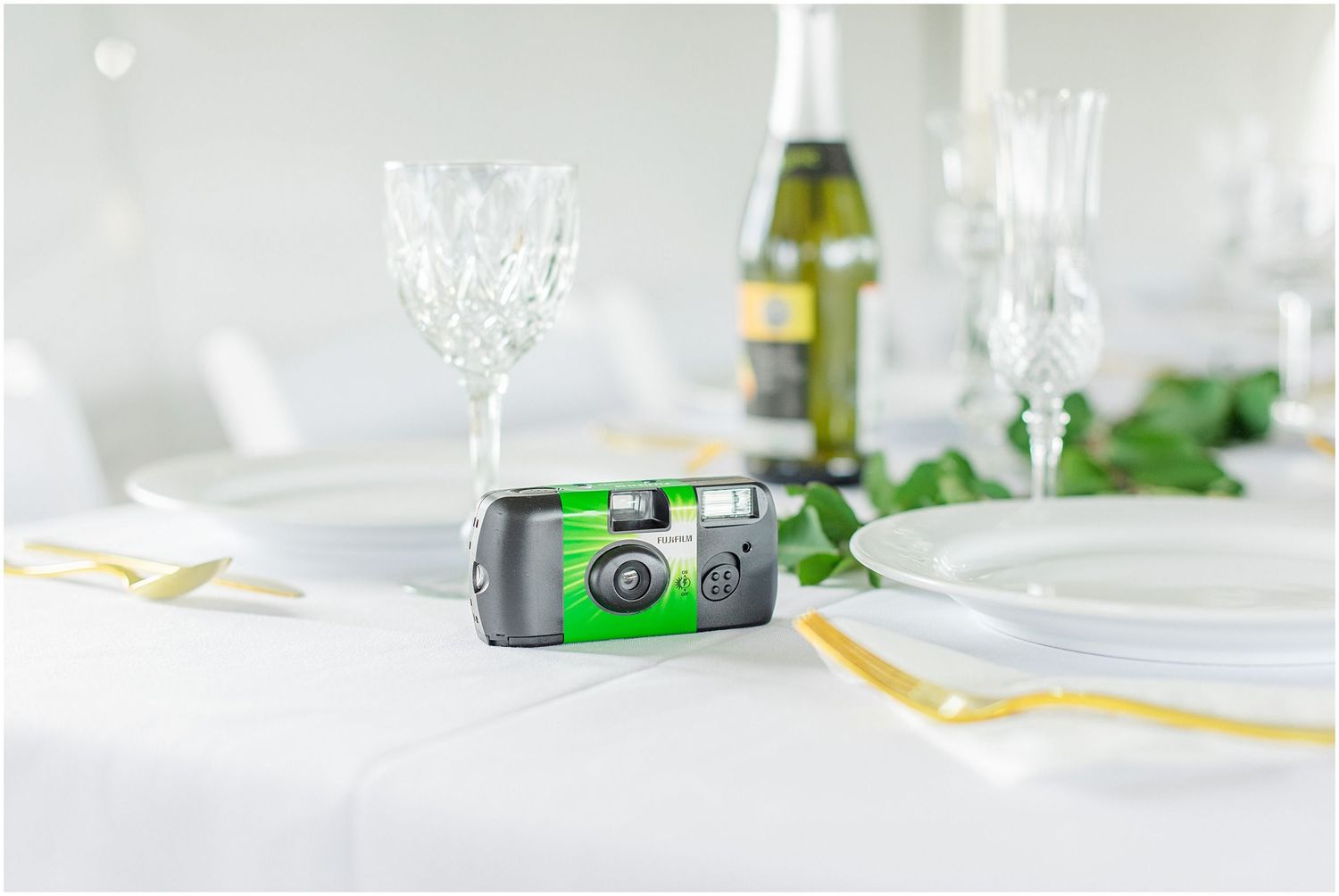 Disposable green film camera on a white reception table with crystal glasses and champagne bottle.