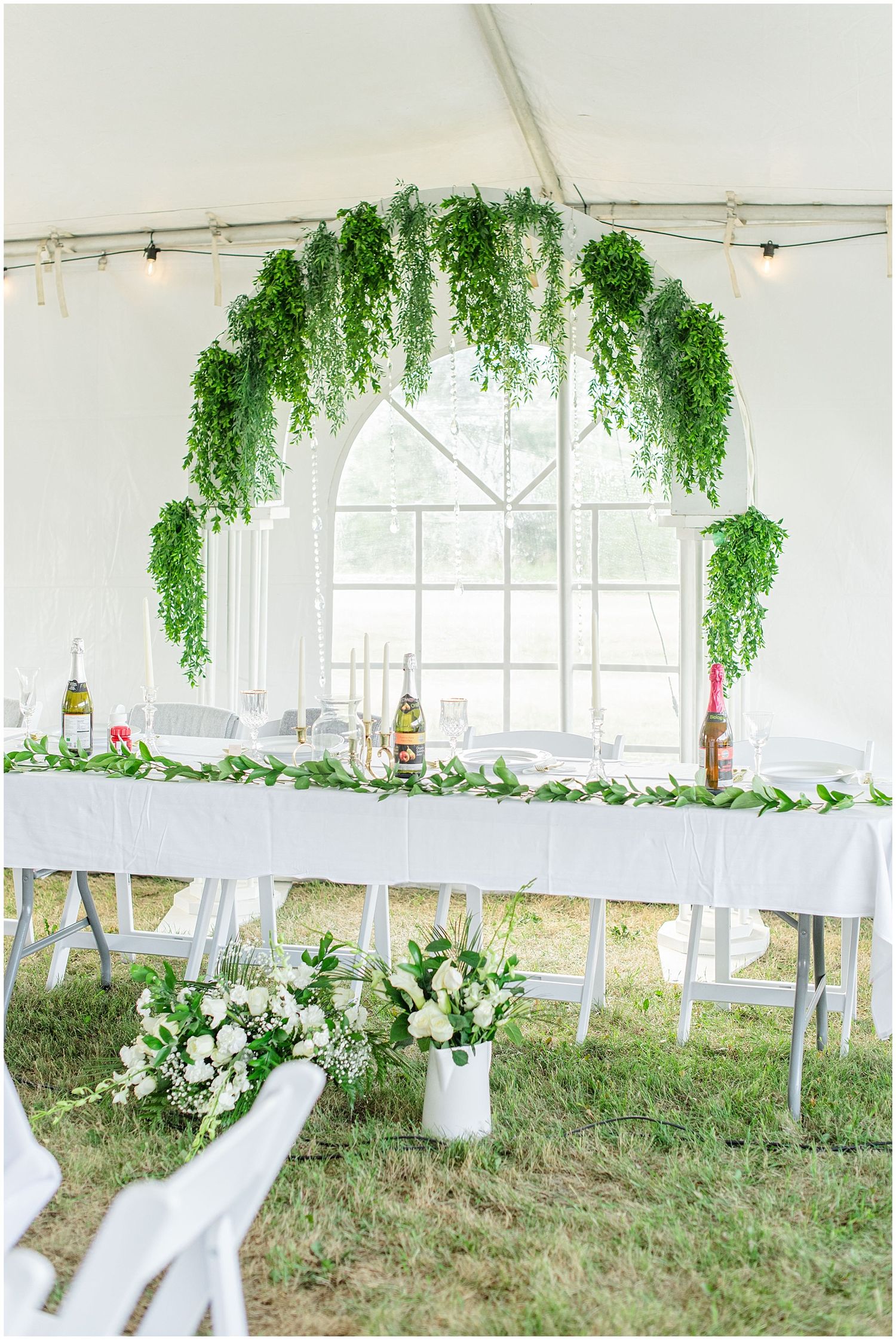 Elegant white tent reception with lush green hanging floral arch over a long white-draped head table.