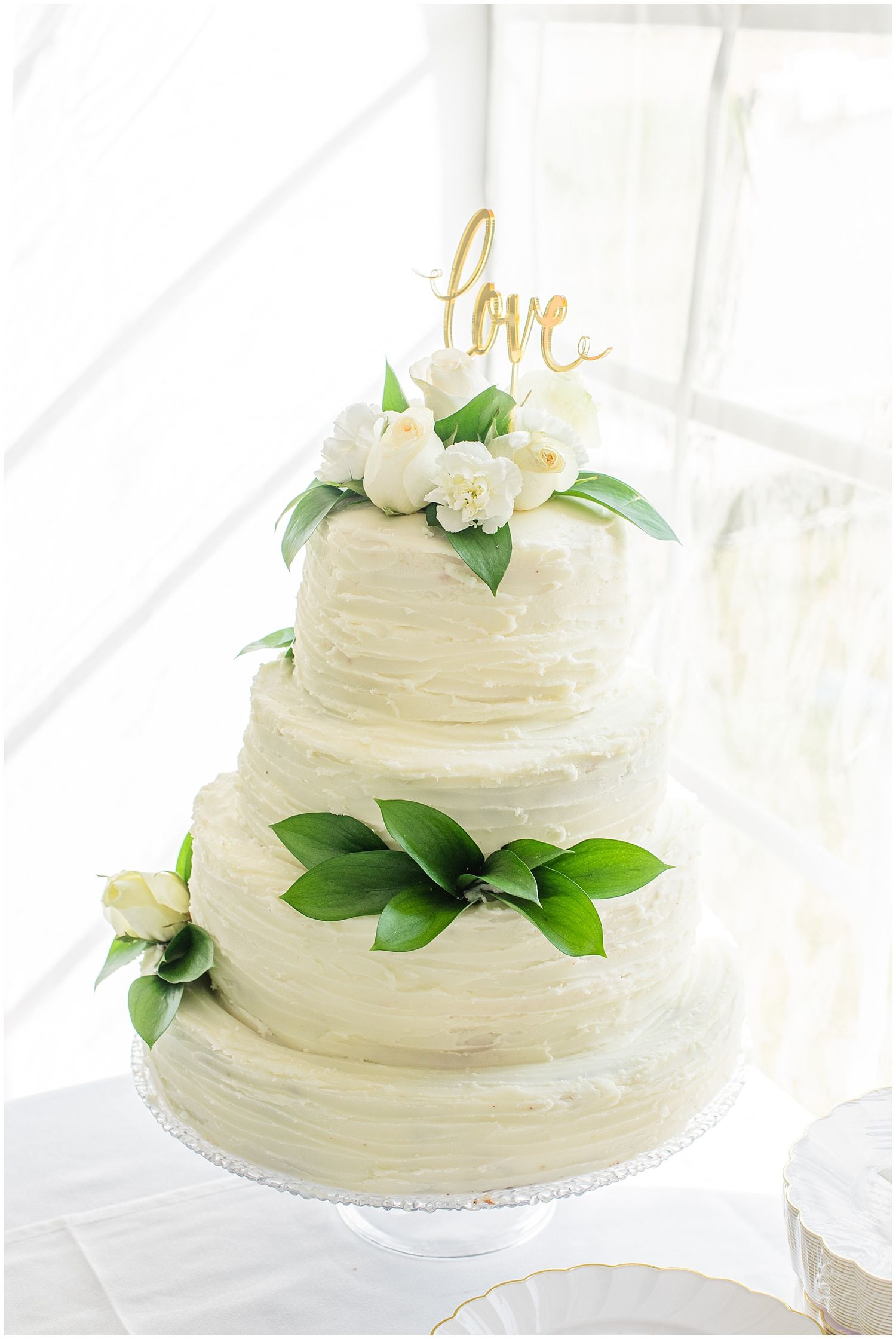 Elegant three-tier white textured wedding cake adorned with green leaves, white roses, and gold love topper.