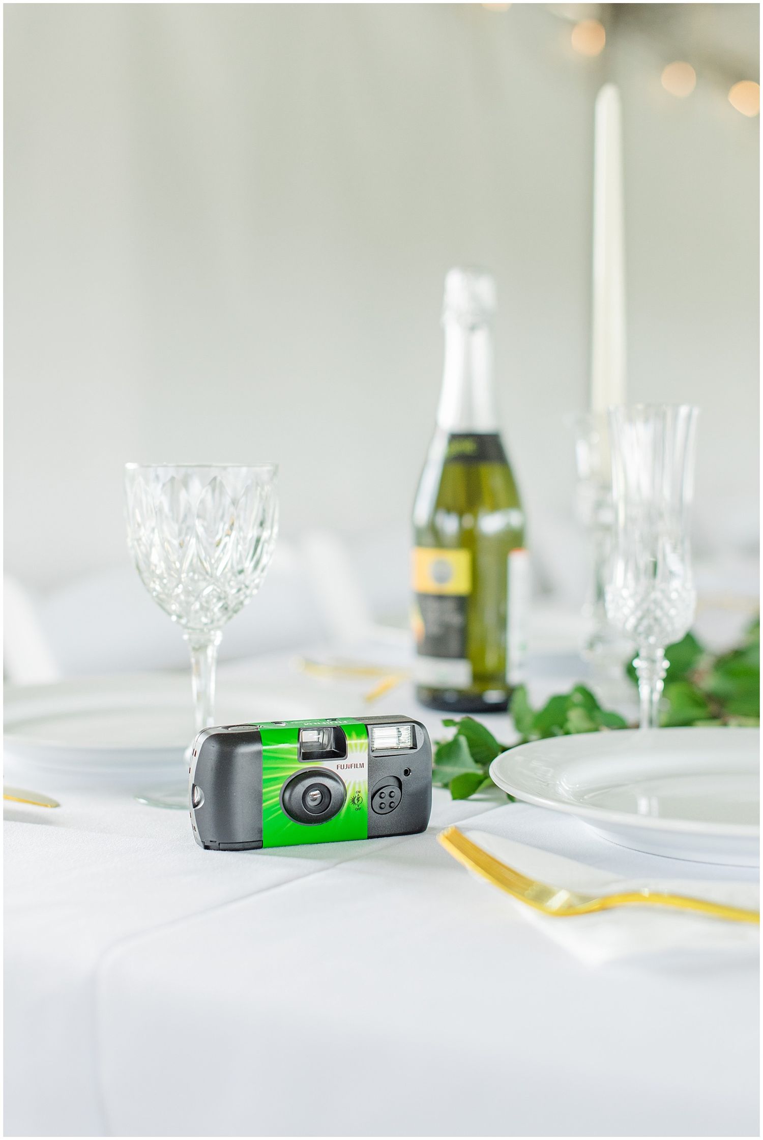 Disposable film camera on white wedding reception table with crystal glasses, champagne bottle, and candles.