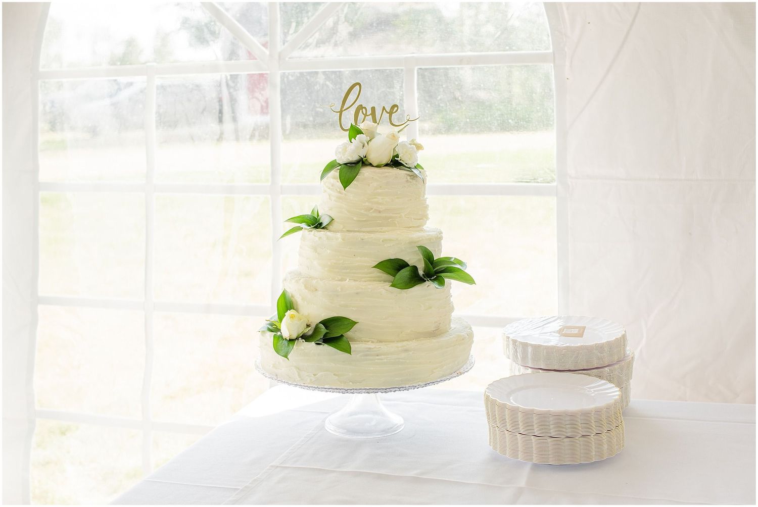 Three-tier white wedding cake with green floral accents and gold love topper displayed on elegant cake stand.