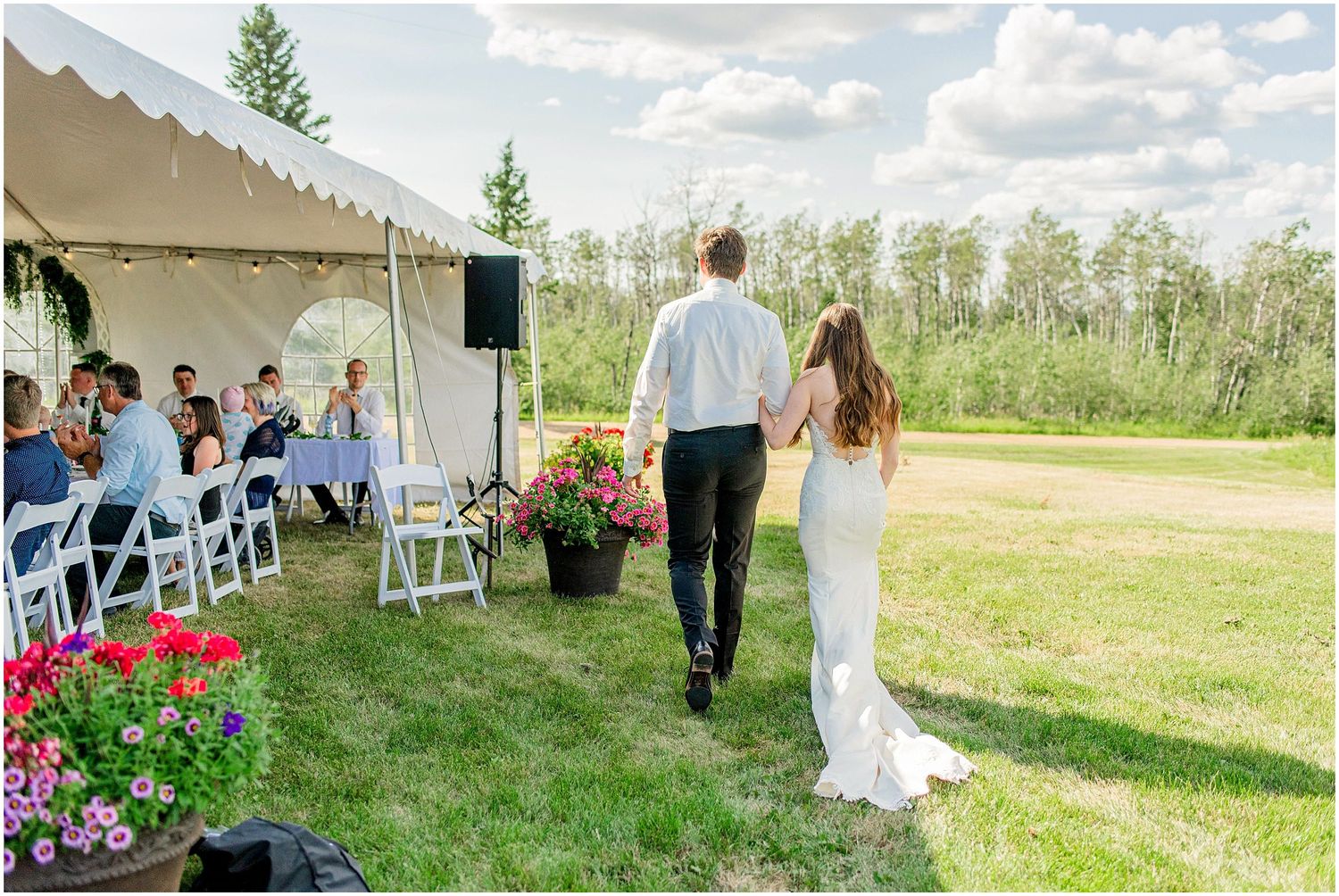 Bride and groom walk hand-in-hand toward wedding reception tent surrounded by colorful flowers and lush greenery.