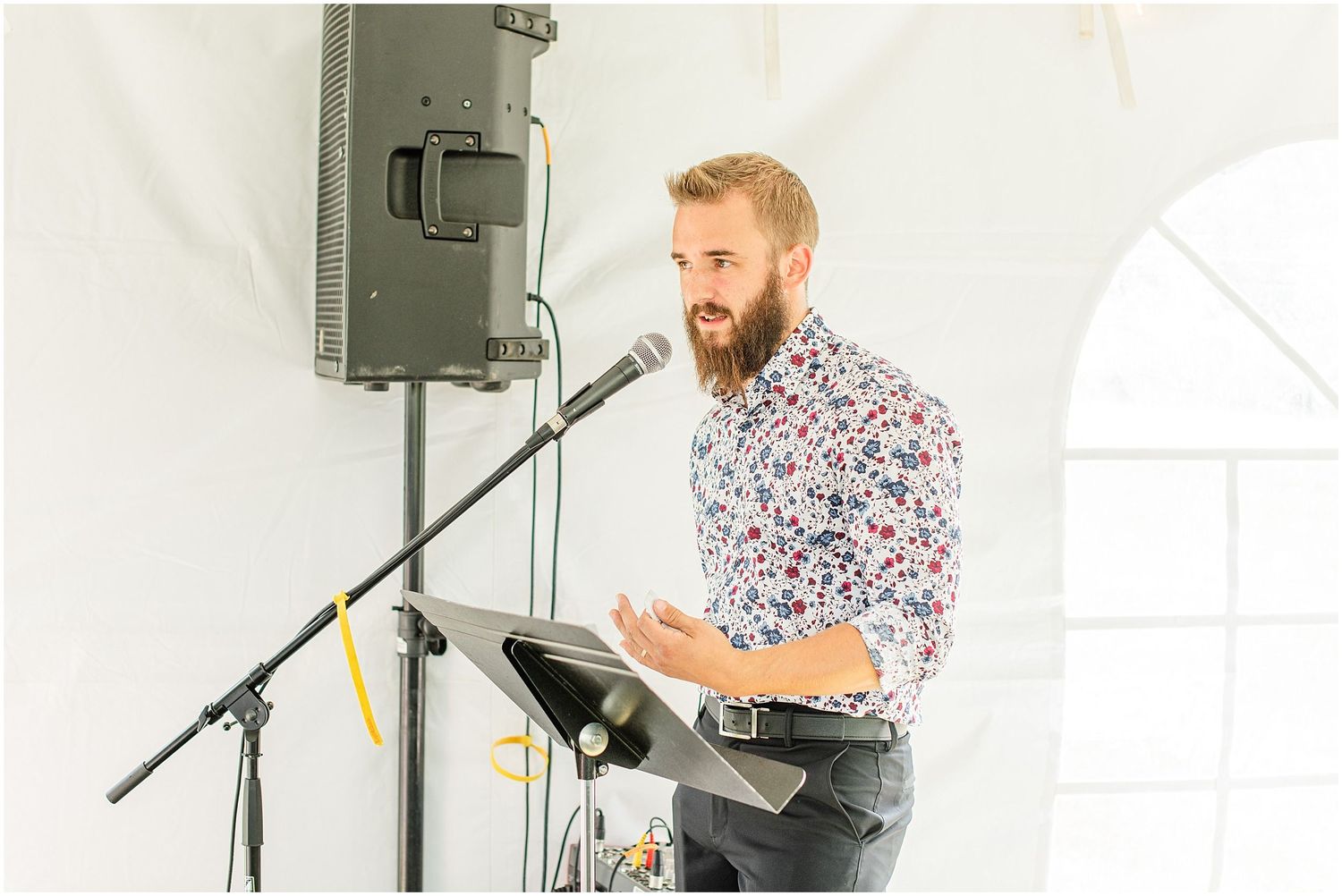 Bearded man in floral shirt speaking at microphone podium during wedding reception under white tent.