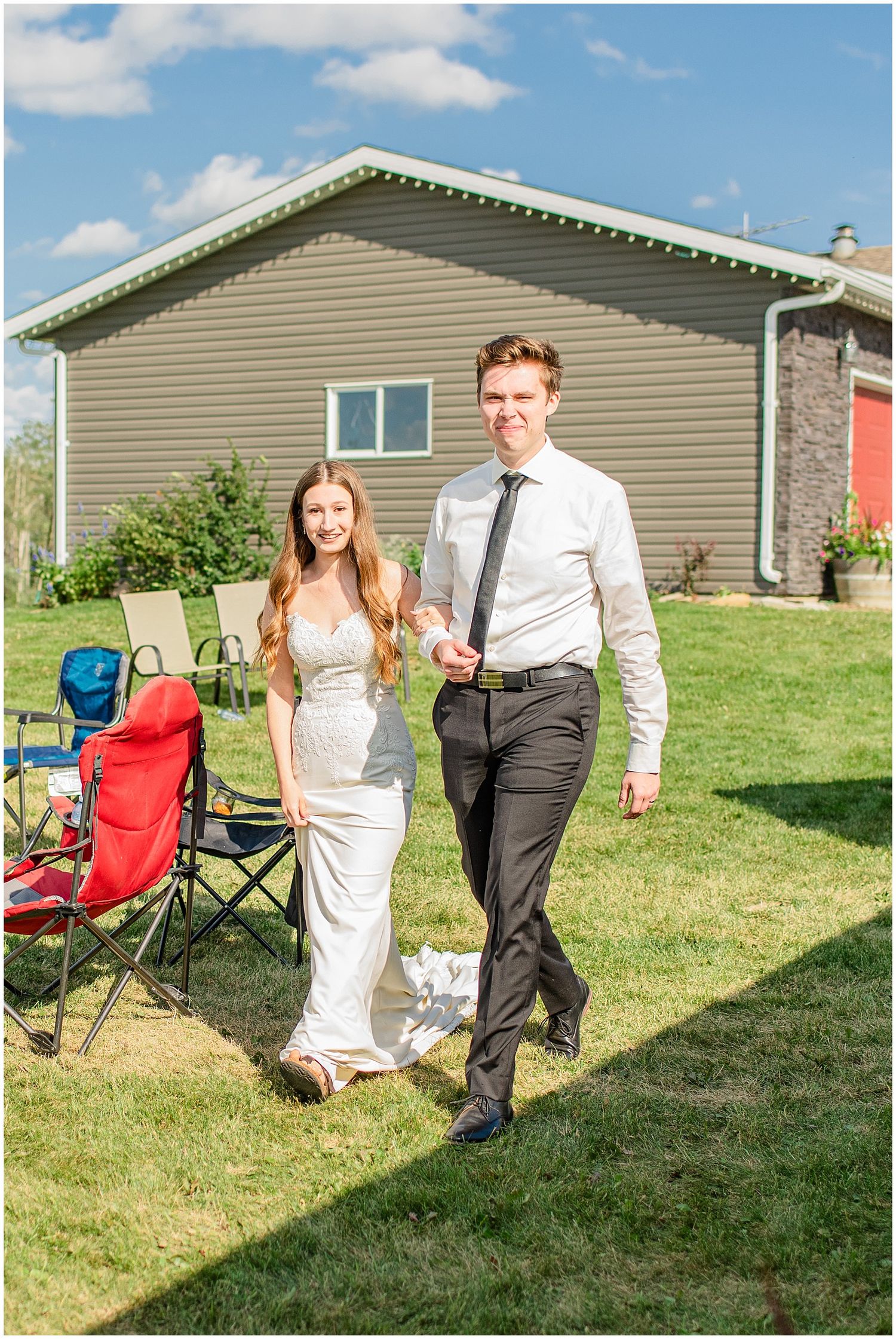 Bride in white gown and groom in black suit walk hand in hand across green lawn at outdoor wedding.
