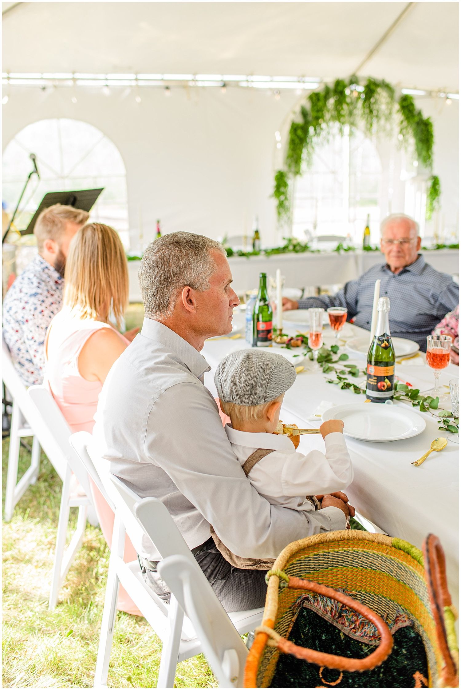 Guests seated at elegant outdoor wedding reception under white tent with greenery decorations and champagne on table.