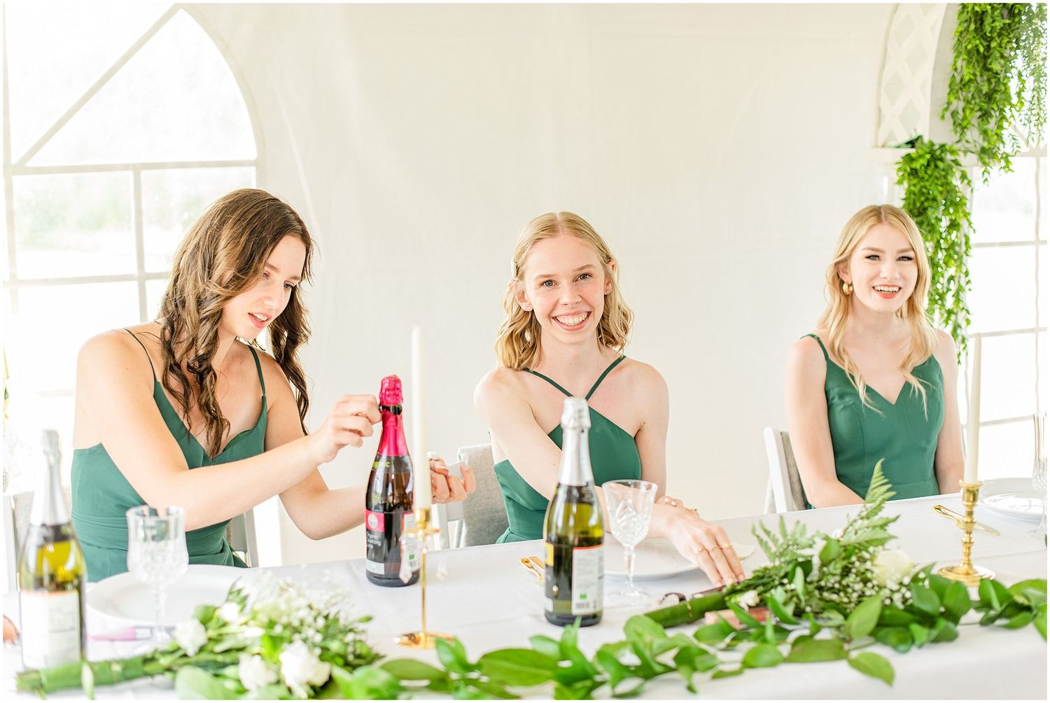 Three bridesmaids in green dresses smile while opening champagne bottles at decorated wedding reception table.