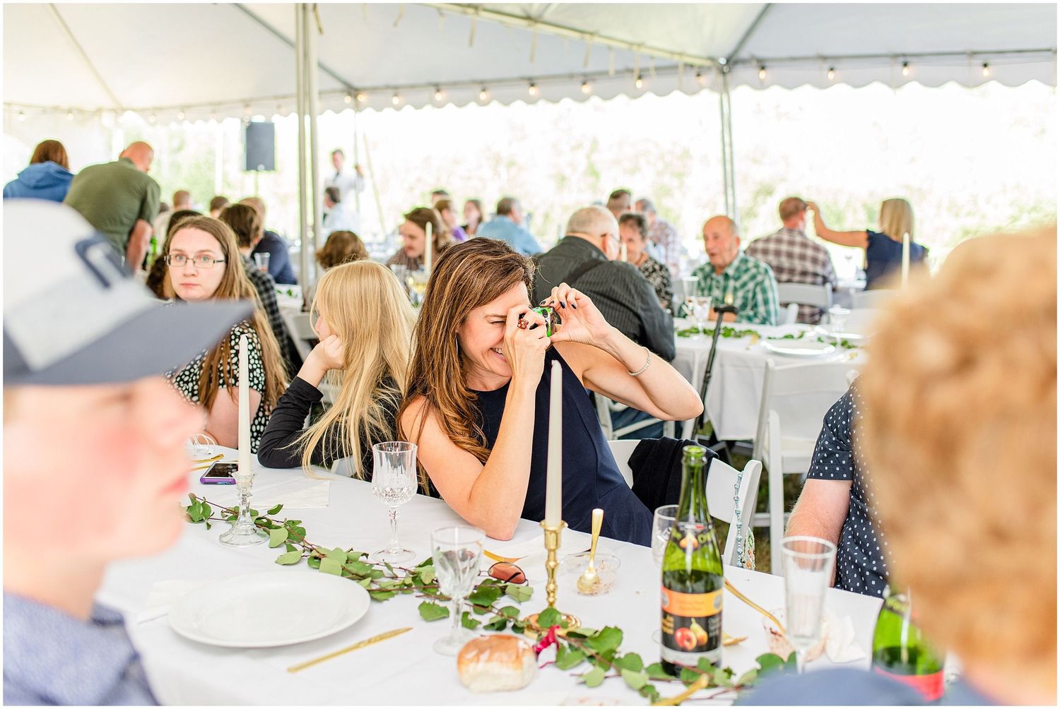 Guests socializing at a wedding reception under a white tent with floral table decor and wine bottles.