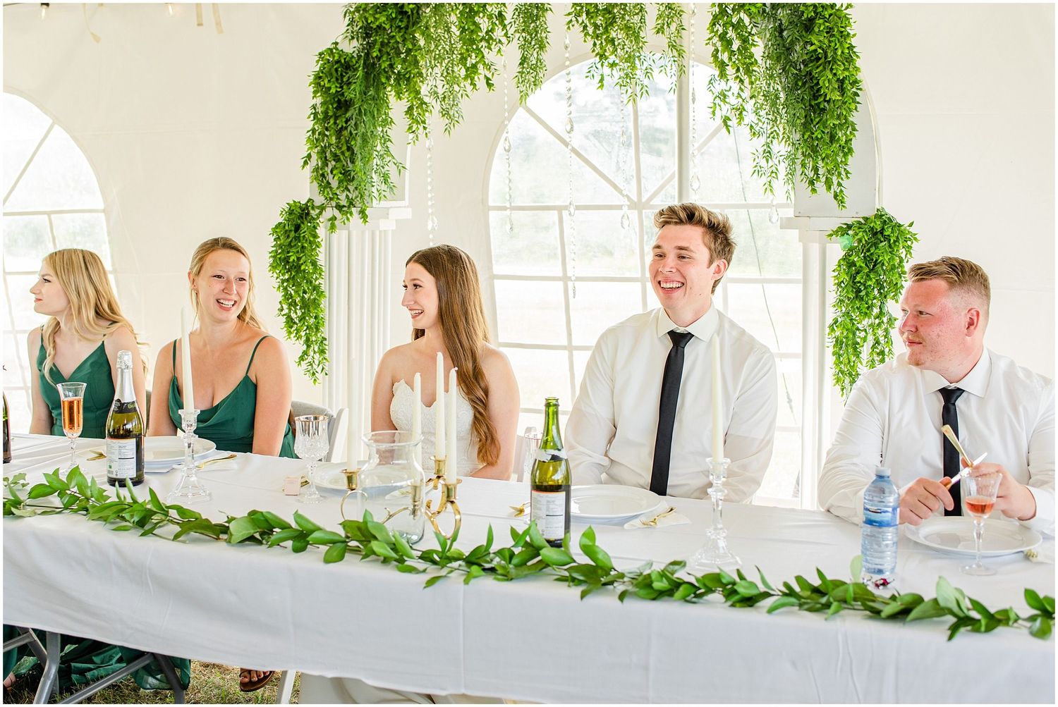 Wedding party laughing at head table decorated with greenery garlands under a white tent.