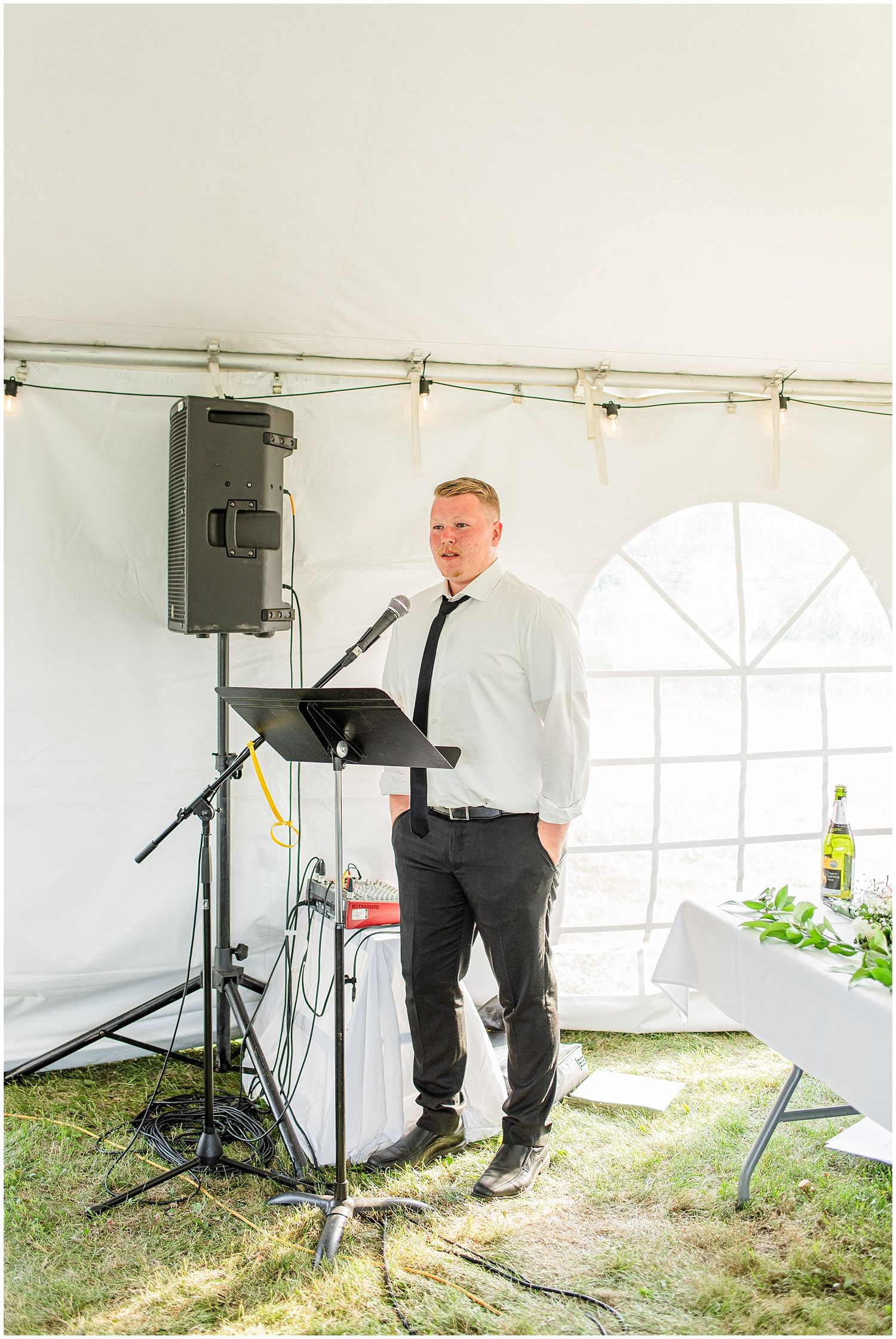 Groomsman in white shirt and black tie delivers speech at microphone podium inside white wedding tent.