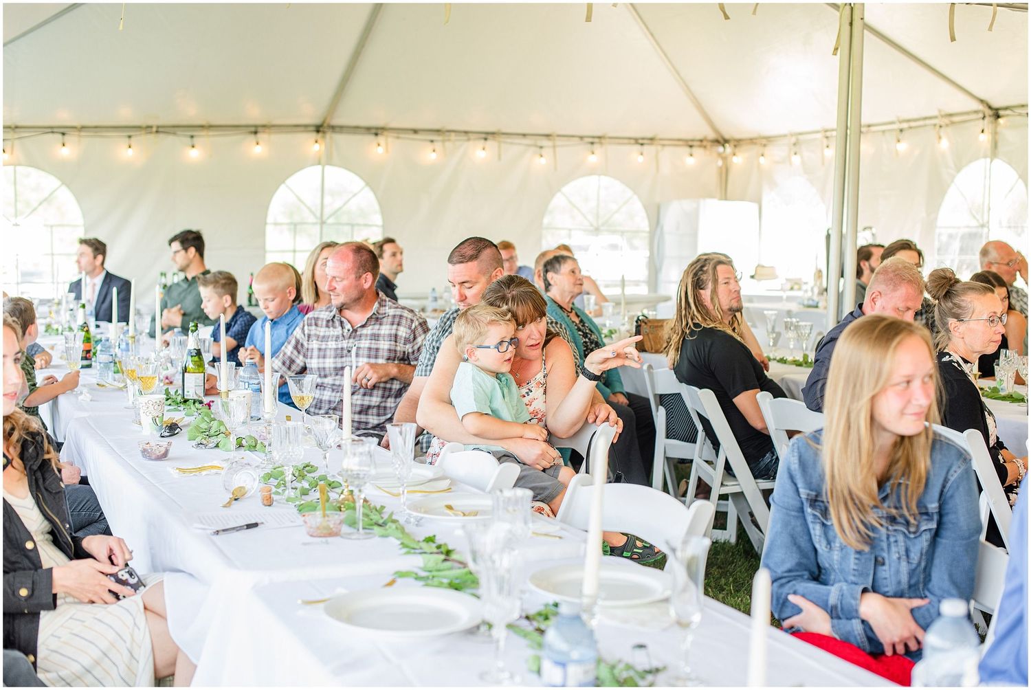 Wedding guests seated at long white-draped tables under a white tent during an outdoor reception celebration.