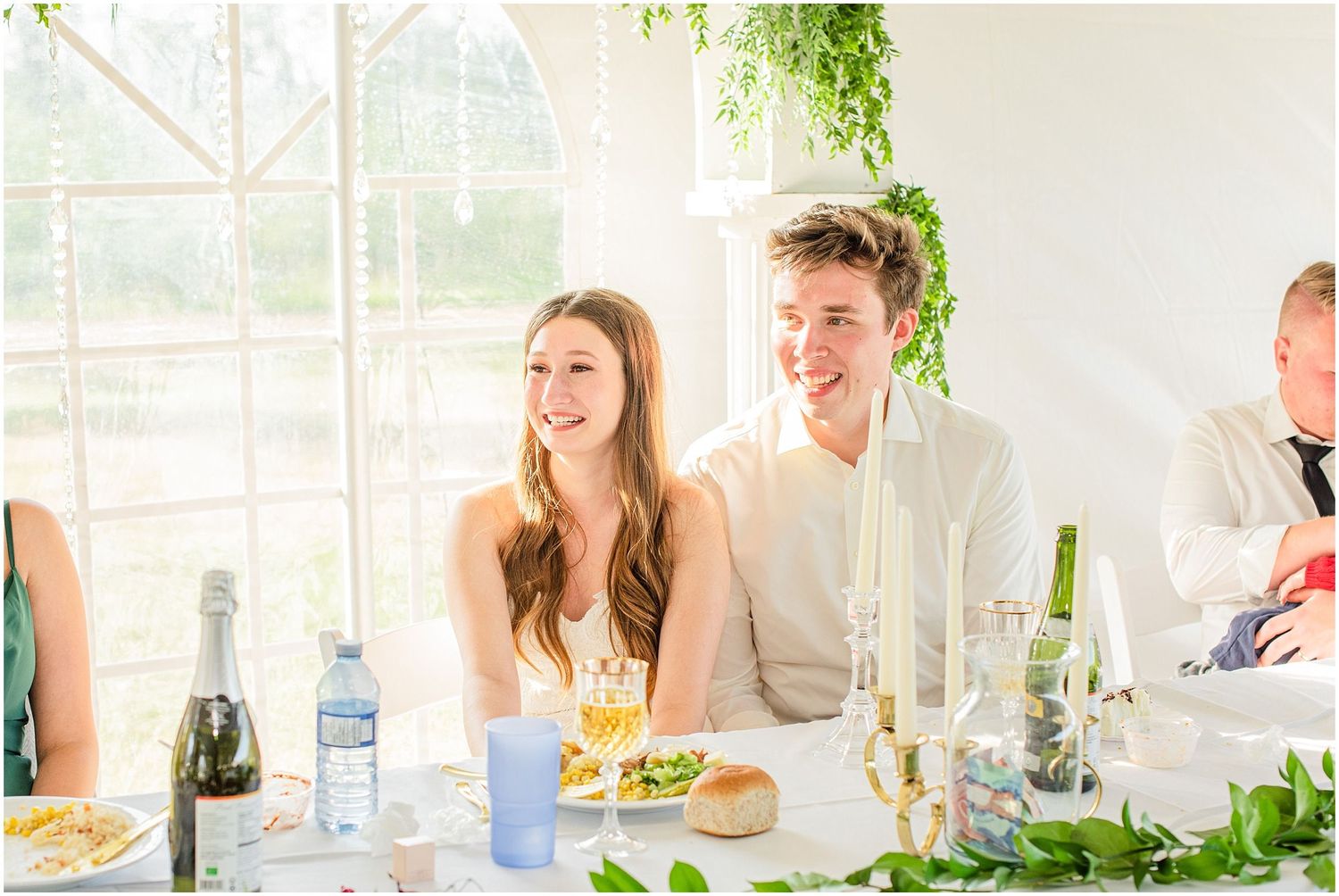 Bride and groom laughing together at wedding reception dinner table with greenery decor.
