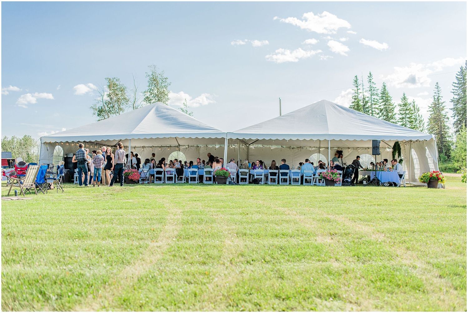Guests gathered under white pole tent for outdoor wedding reception on lush green grass on a sunny day.