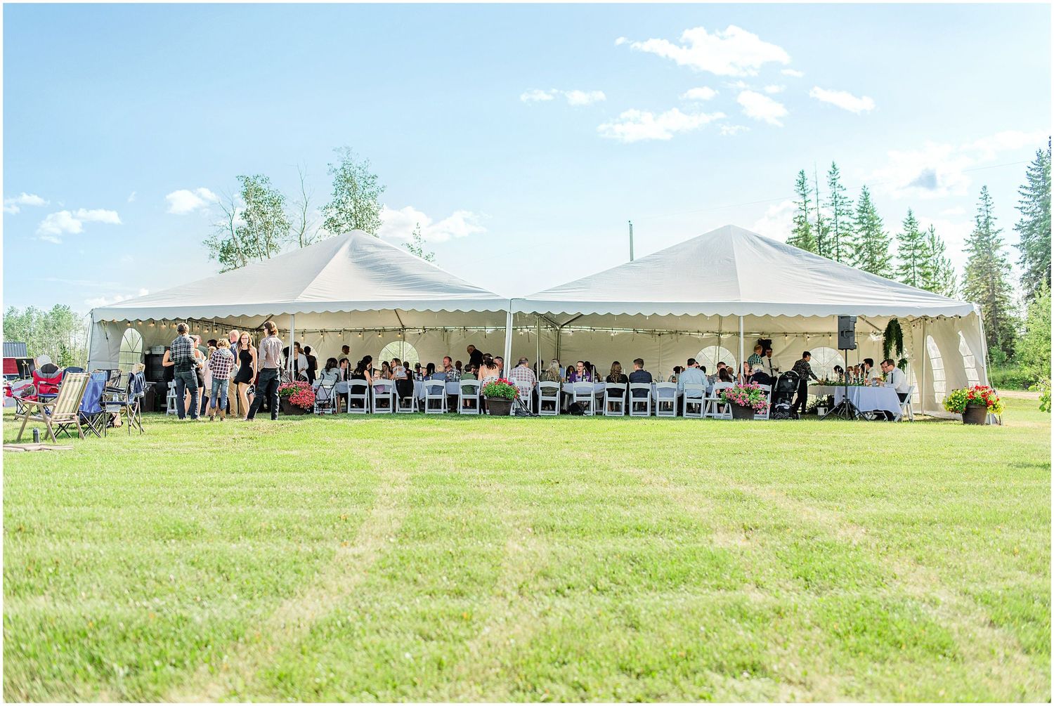 Outdoor wedding reception under large white tent on green lawn surrounded by tall trees on sunny day.
