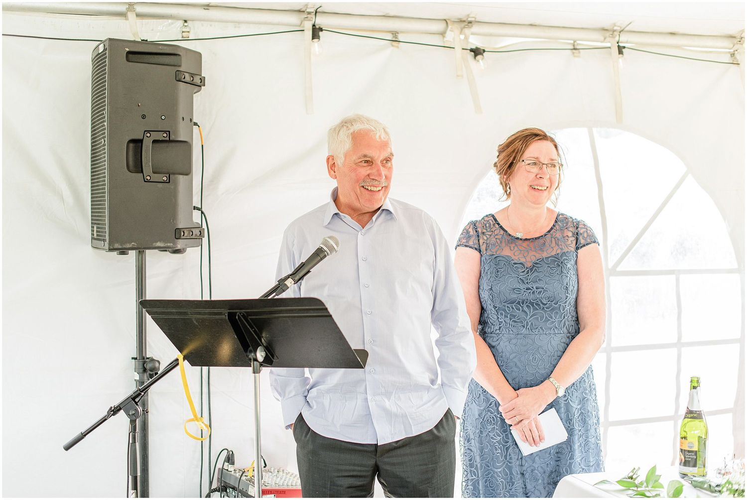 Man and woman smiling while giving speech at wedding reception with microphone and music stand inside tent.