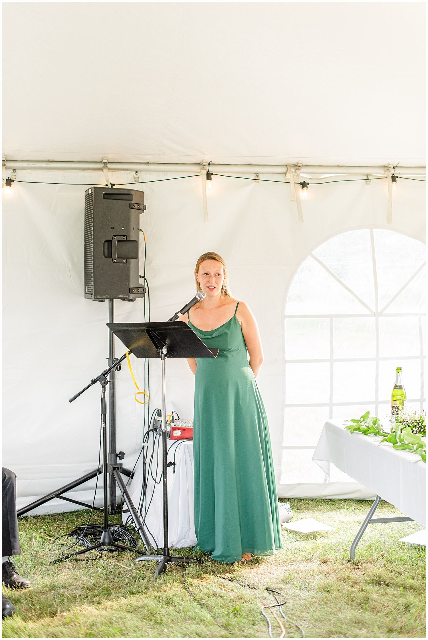 Woman in green gown speaks at microphone during outdoor wedding reception under white tent.